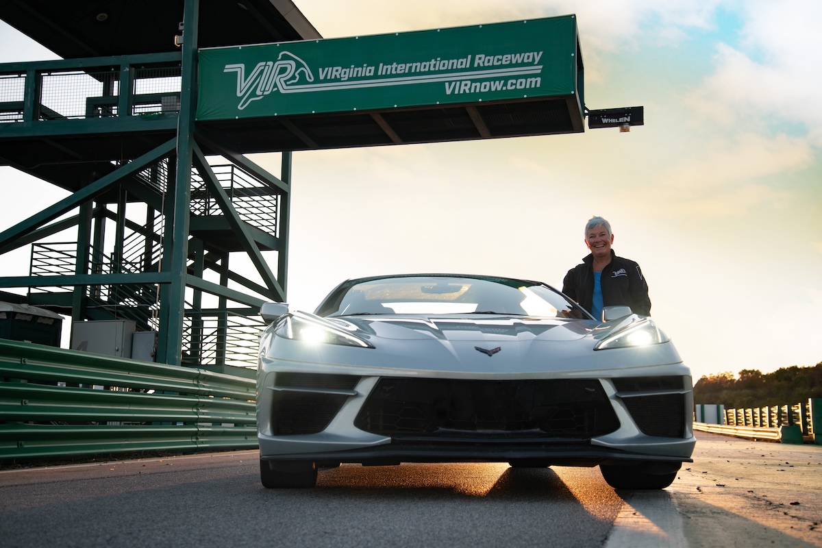 A person stands next to a silver sports car on a racetrack under a green Virginia International Raceway sign at sunset, embodying the spirit of Raceway Queen Connie Nyholm’s legendary track.