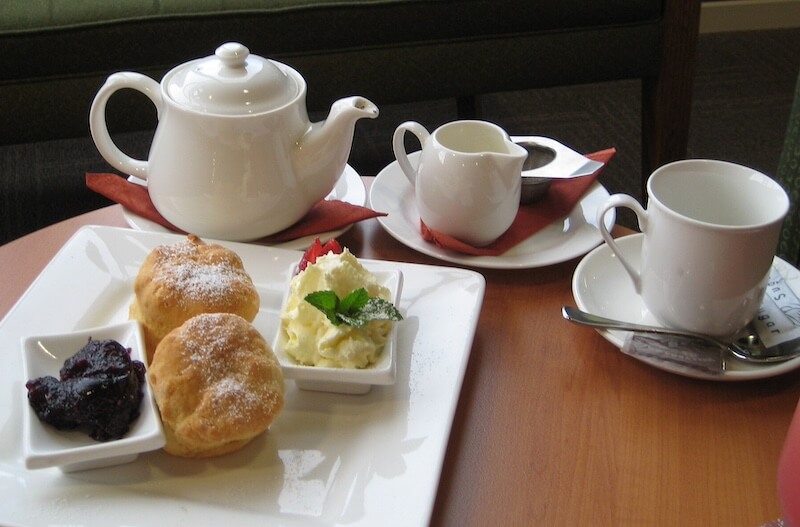 10 More Britishisms We Love - 2 A tea set with a teapot, milk jug, and cup is arranged beside a plate holding two scones, clotted cream, and jam—a classic spread on a wooden table.