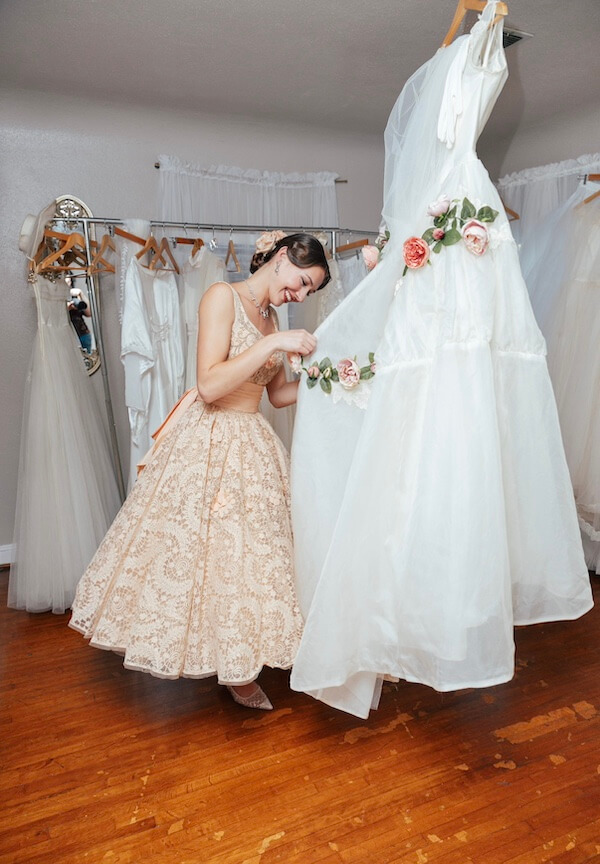 Estelle Amore: New Vintage Bridal Destination - 4 A woman in a beige lace dress adjusts an Estelle Amore white wedding gown decorated with flowers in a bridal boutique.