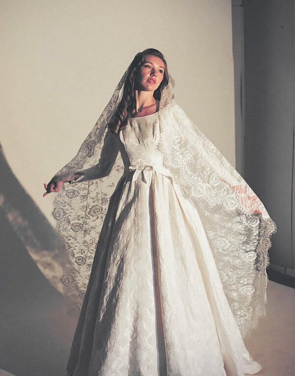 Estelle Amore: New Vintage Bridal Destination - 5 Estelle Amore shines in a long white wedding dress with a lace veil, standing against a plain background and looking slightly to the side.