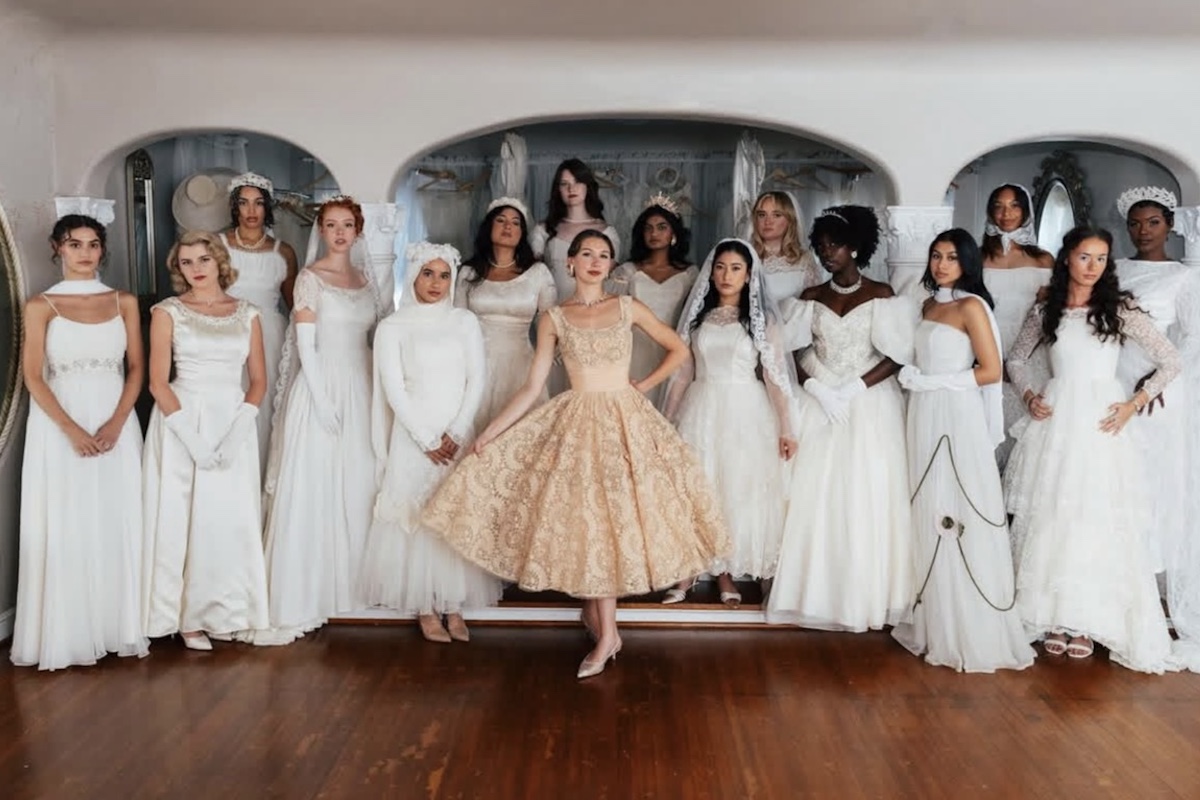 A group of women in white bridal dresses stand in a row indoors, with one woman in the center wearing a short, gold dress by Estelle Amore.
