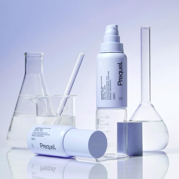 12 In-Flight Plane Accessories You Need - 10 Two white Prequel skincare bottles, perfect as plane accessories, are placed among clear laboratory glassware filled with water on a reflective surface with a light purple background.