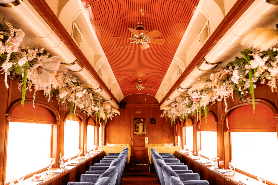Elegant vintage train dining car with blue velvet seats, wooden paneling, large windows, and floral arrangements above the windows—perfect for savoring luxury train journeys in timeless style.