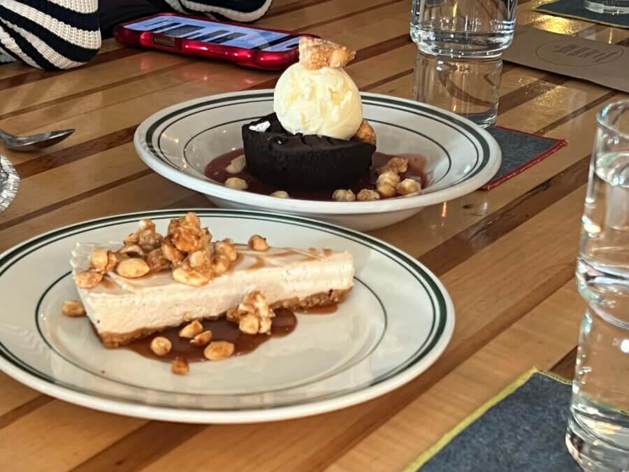Your Guide to Chattanooga's Culinary Scene - 3 Two dessert plates on a wooden table in Chattanooga: one with cheesecake topped with nuts and caramel, another with chocolate cake, ice cream, and nuts. Glasses of water and a phone are nearby.