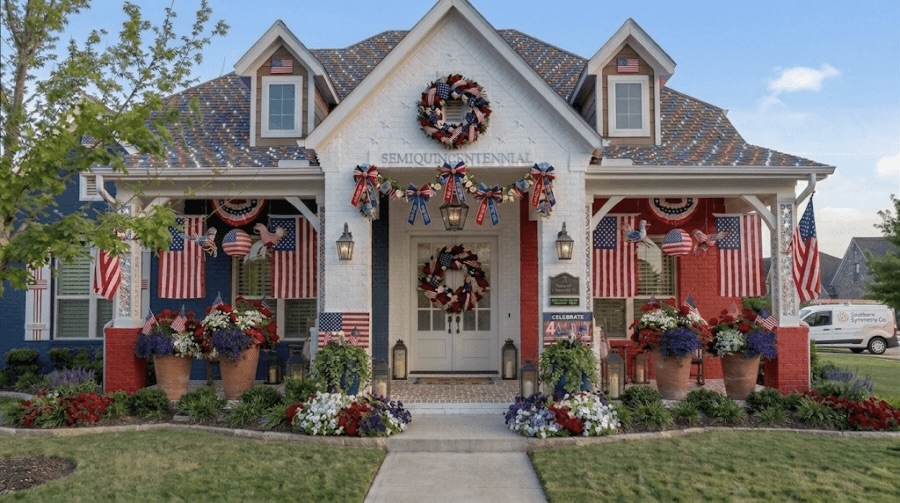 A house decorated with American flags, red, white, and blue banners, floral arrangements, and patriotic wreaths creates charming front porch decor for a festive look on the porch and windows.