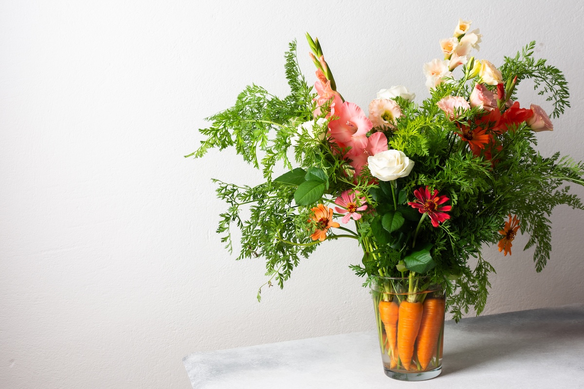 A clear glass vase holds a bouquet of assorted flowers and greenery, with whole carrots submerged in water at the base—a charming centerpiece to elevate your Easter spread.