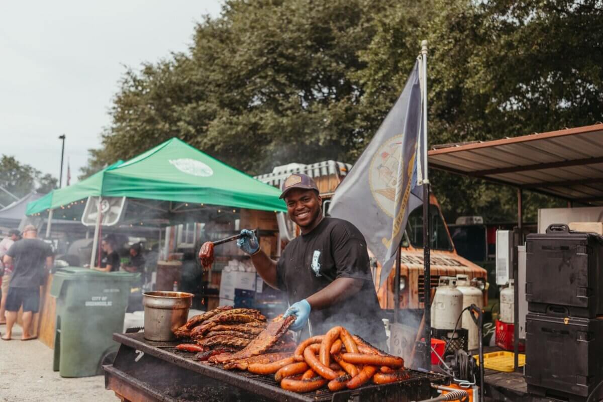 A man grilling sausages and ribs at an outdoor food stand, holding tongs and smiling, with tents and trees in the background—capturing the lively spirit found at some of the best festivals in the South.