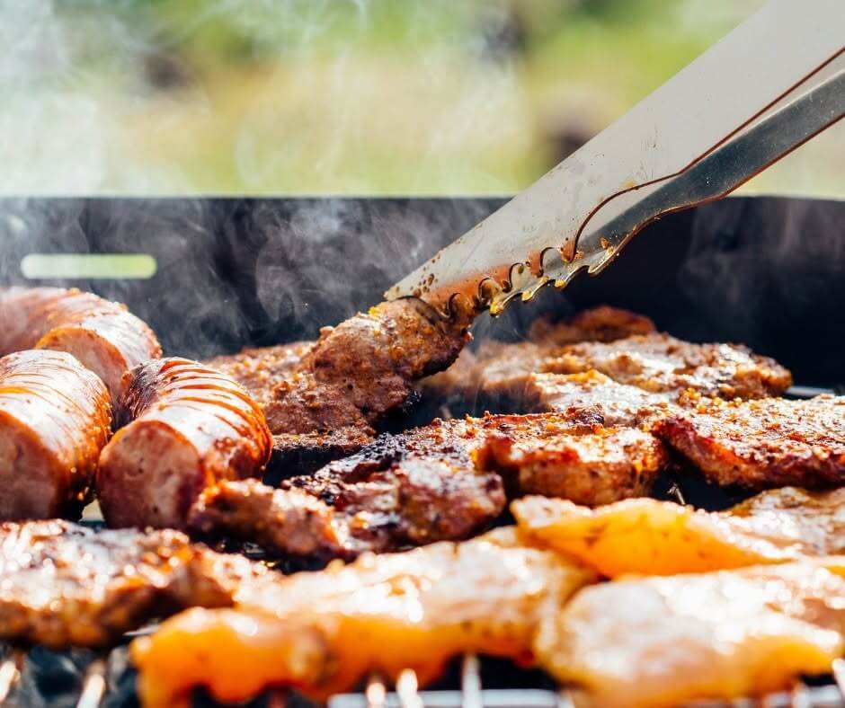 Close-up of various meats, including sausages and steaks, grilling on a barbecue with tongs picking up a piece, and smoke rising from the food—a mouthwatering scene reminiscent of the flavorful feasts found at the best festivals in the South.