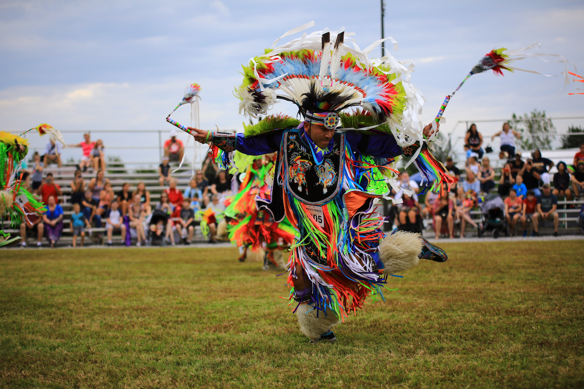 A person in colorful traditional regalia dances at an outdoor event—one of the best festivals in the South—with spectators seated on bleachers in the background.