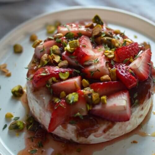 RECIPE: Strawberry-Thyme Baked Brie - 3 A round wheel of cheese topped with sliced strawberries, chopped pistachios, herbs, and honey on a white plate.