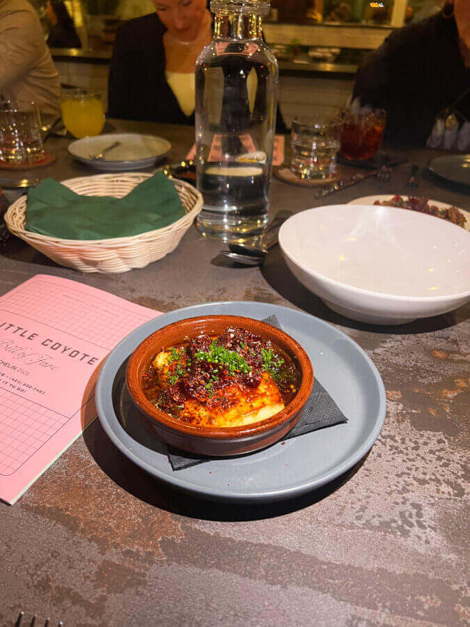 Your Guide to Chattanooga's Culinary Scene - 11 A small dish of baked cheese topped with herbs and sauce sits on a gray plate in a Chattanooga restaurant, surrounded by a water bottle, menu, and other dinnerware for a cozy dining experience.