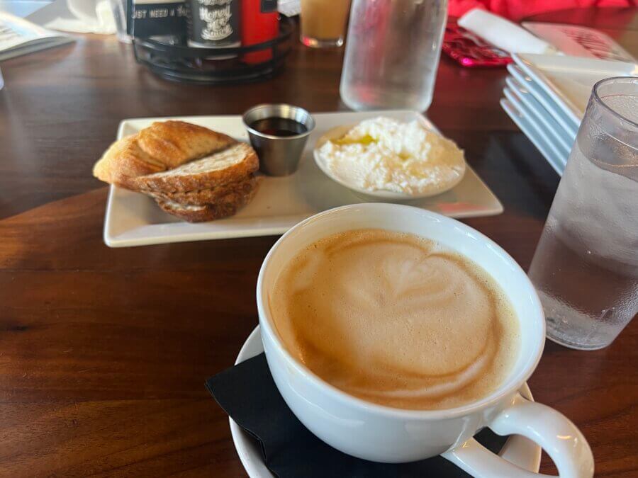 Your Guide to Chattanooga's Culinary Scene - 10 A cup of latte on a saucer, beside a plate with a croissant, a metal cup of syrup, and whipped ricotta—served on a wooden table in the heart of Chattanooga.