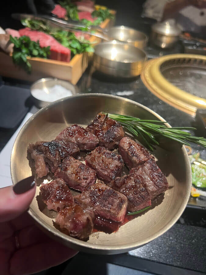 Your Guide to Chattanooga's Culinary Scene - 9 A hand holds a bowl of grilled beef cubes garnished with a sprig of rosemary, with side dishes and a grill visible in the background, capturing the flavors of Chattanooga cuisine.