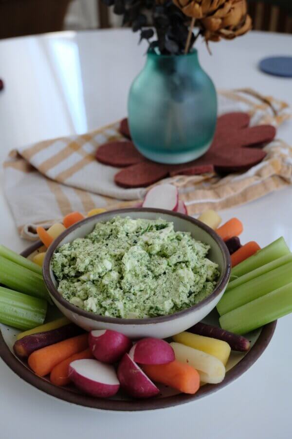 3 Quick and Easy Appetizers for Spring - 2 A bowl of green vegetable dip is surrounded by celery, radishes, and multicolored carrots on a plate, with a vase and napkin in the background.