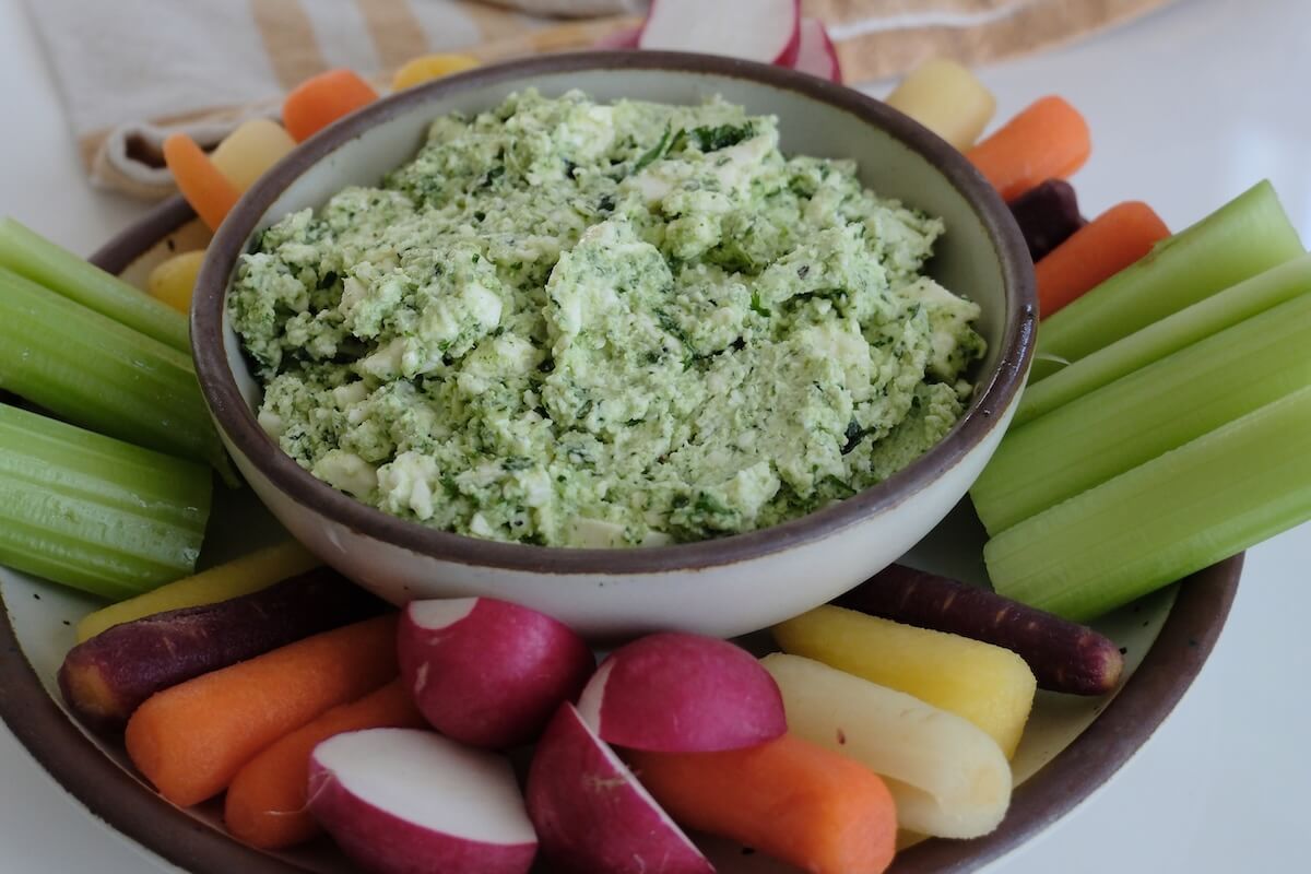 A bowl of herbed cheese dip is surrounded by celery, carrots, and radishes arranged on a plate.