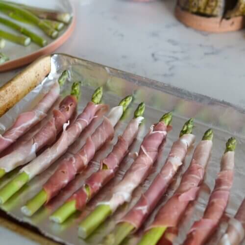 RECIPE: Easy Prosciutto-Wrapped Asparagus - 3 Asparagus spears wrapped in prosciutto are arranged on a foil-lined baking sheet, ready to be cooked; trimmed asparagus is visible on a plate in the background.
