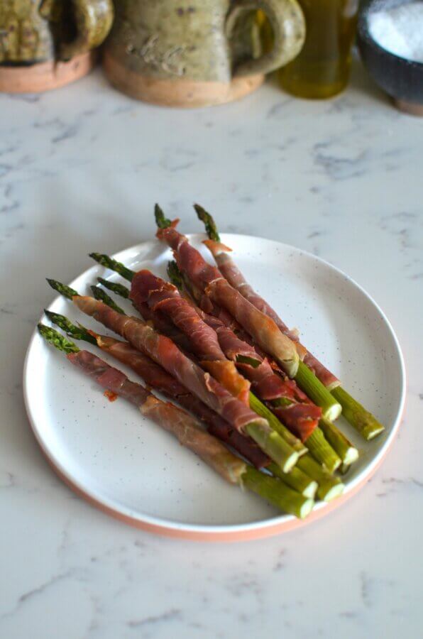 3 Quick and Easy Appetizers for Spring - 4 A white plate with several asparagus spears wrapped in prosciutto, placed on a marble countertop.