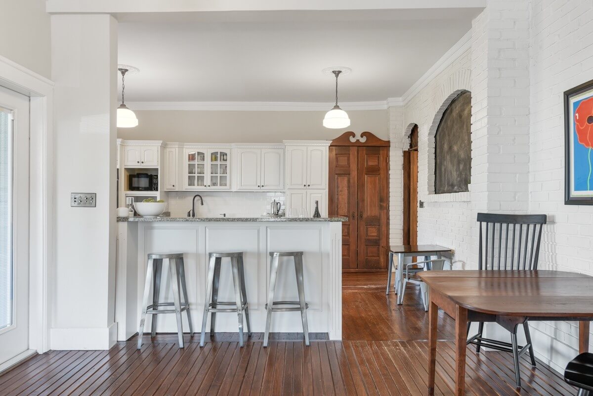 Modern kitchen at 903 Russell St, Nashville, with white cabinets, metal bar stools at the breakfast bar, wooden floors, and a dining table. A wooden door and white brick walls complete this inviting Nashville space.