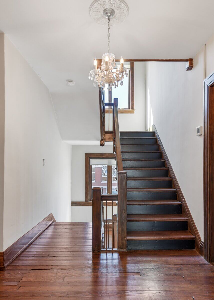 A wooden staircase with dark steps and a wooden railing leads to an upper floor at 903 Russell St, Nashville, with a chandelier above and natural light streaming through the windows.