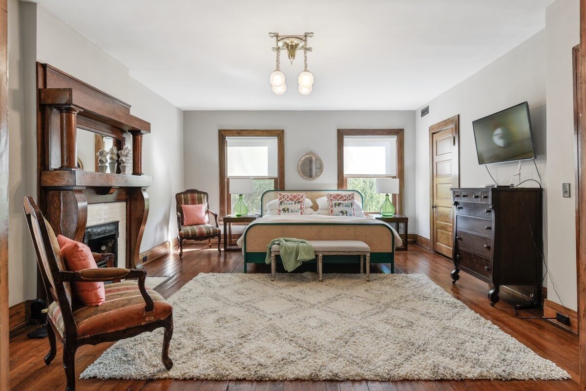 Spacious bedroom at 903 Russell St, Nashville, featuring a bed, two windows, wall-mounted TV, fireplace, wooden furniture, and a large cream-patterned rug on a wooden floor.