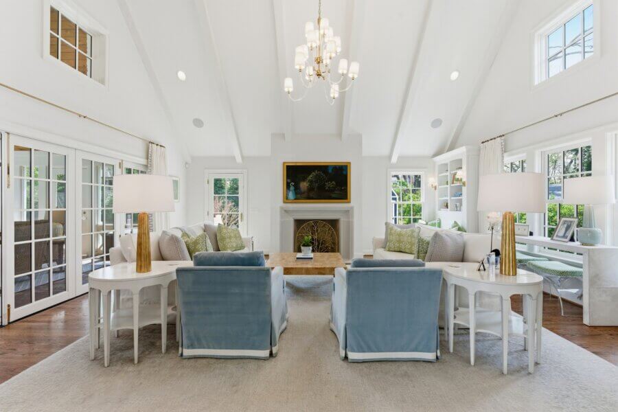 Bright living room with vaulted ceiling, chandelier, fireplace, white sofas, blue armchairs, and large windows. Glass doors overlook a lush garden on Sperry Ave in Nashville.