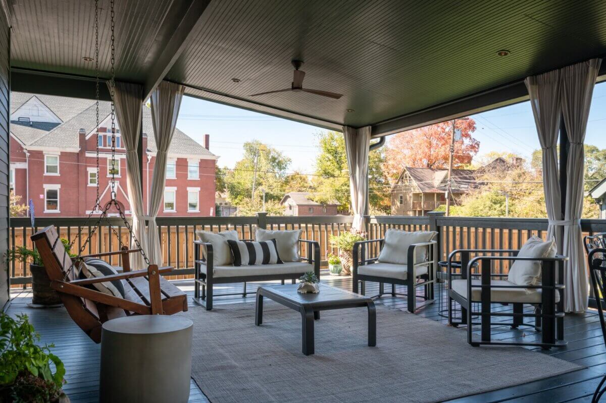 Relax on this covered outdoor patio at 903 Russell St, Nashville, featuring cushioned seating, a swing, coffee table, and rug. Curtains frame the corners while trees and city views complete the inviting atmosphere.