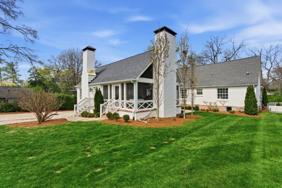 Charming single-story white brick house on 3619 Sperry Ave, Nashville TN, with a screened porch, two chimneys, manicured lawn, young trees, and a clear blue sky in the background.