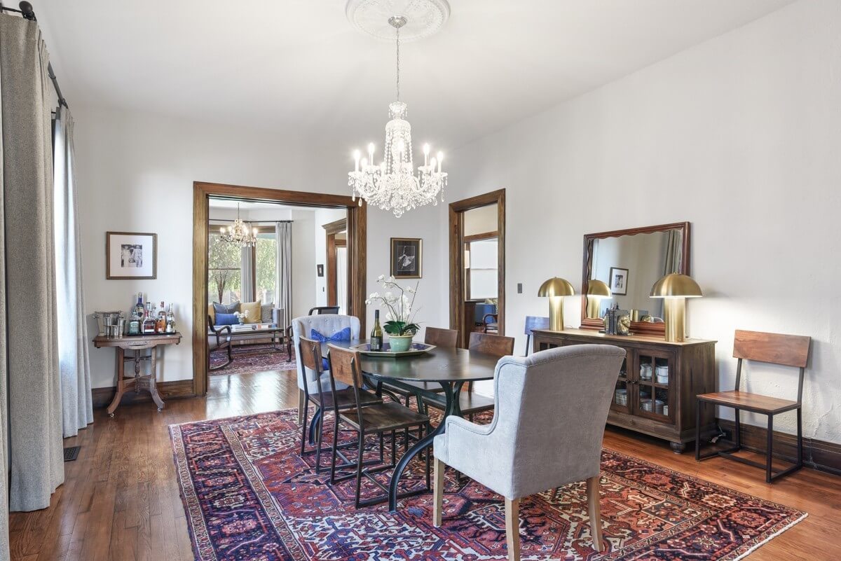 A dining room at 903 Russell St, Nashville features a dark wood table with mixed chairs, a chandelier, sideboard with lamps, and a red patterned rug. An adjoining living room is visible through an open doorway.