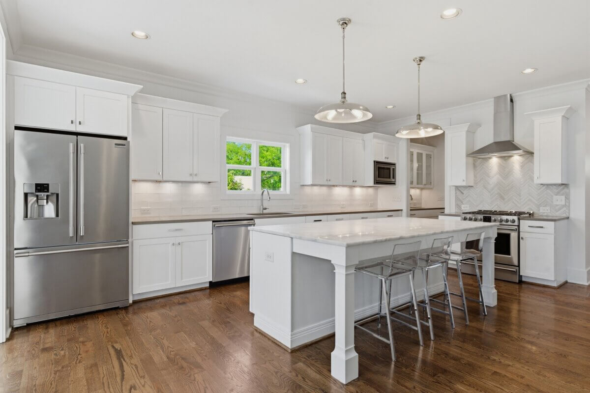 Modern kitchen in Nashville TN featuring white cabinets, stainless steel appliances, a marble island with three clear bar stools, pendant lights, and hardwood floors.