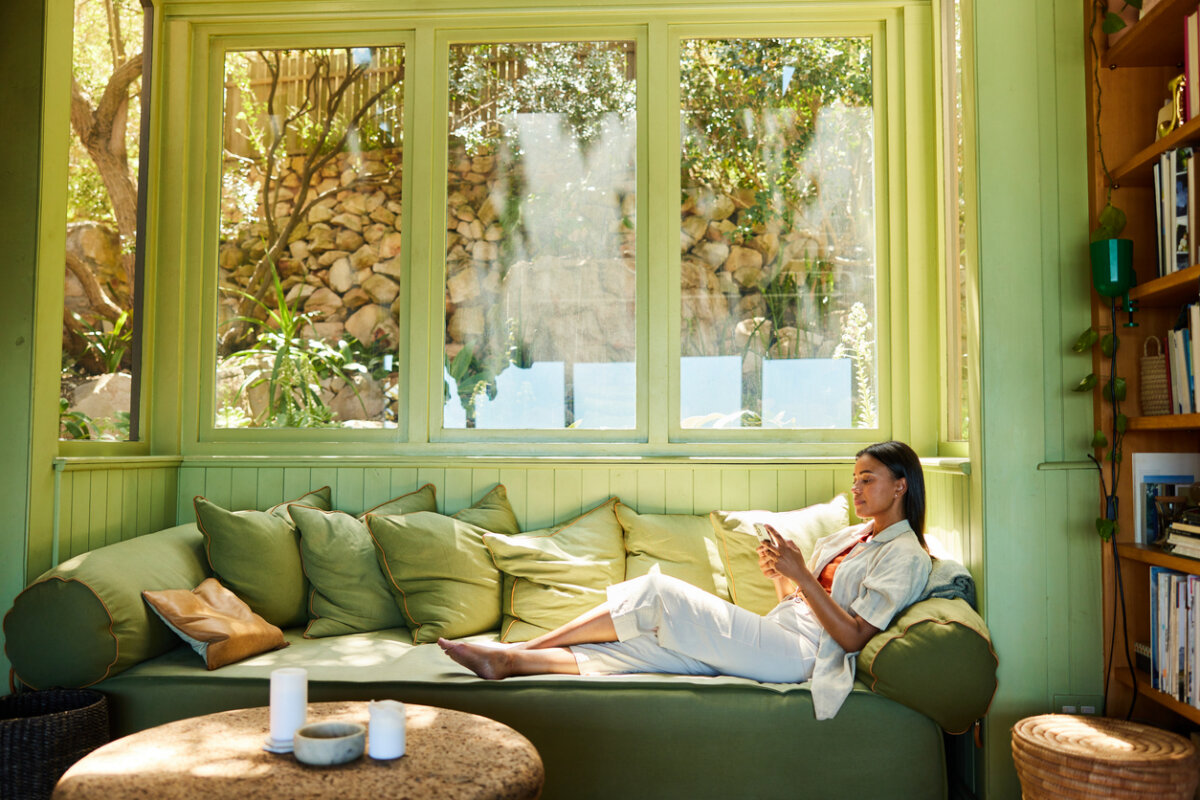 A person sits on a green sofa by large windows, browsing their smartphone in a sunlit, cozy living room with shelves and outdoor views—a perfect space for inspiration from the home design guild.