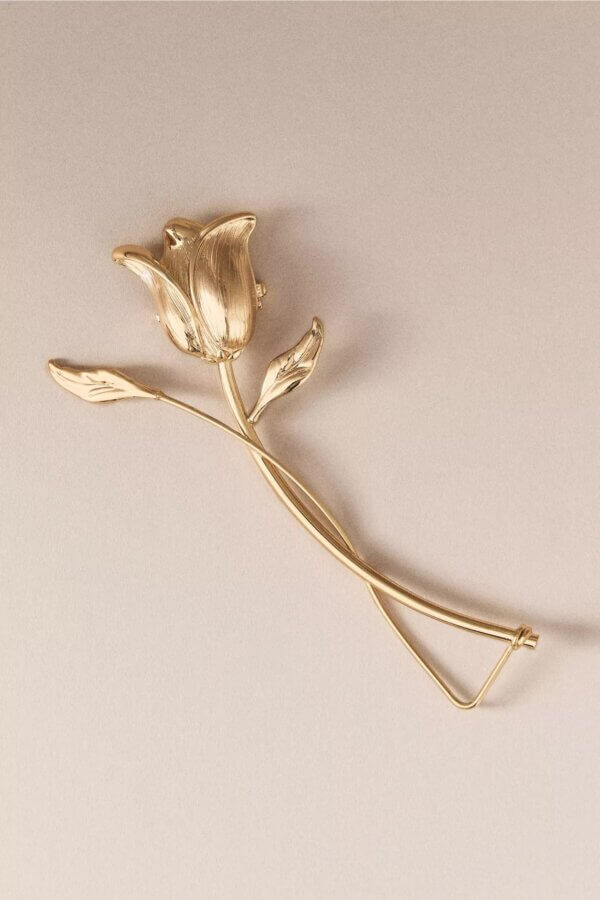 It's the Year of the Brooch! Here's How to Style Them - 6 A gold brooch shaped like a rose with two leaves and a slightly curved stem is displayed on a neutral background, capturing the elegance and charm that defines the Year Of The Brooch.