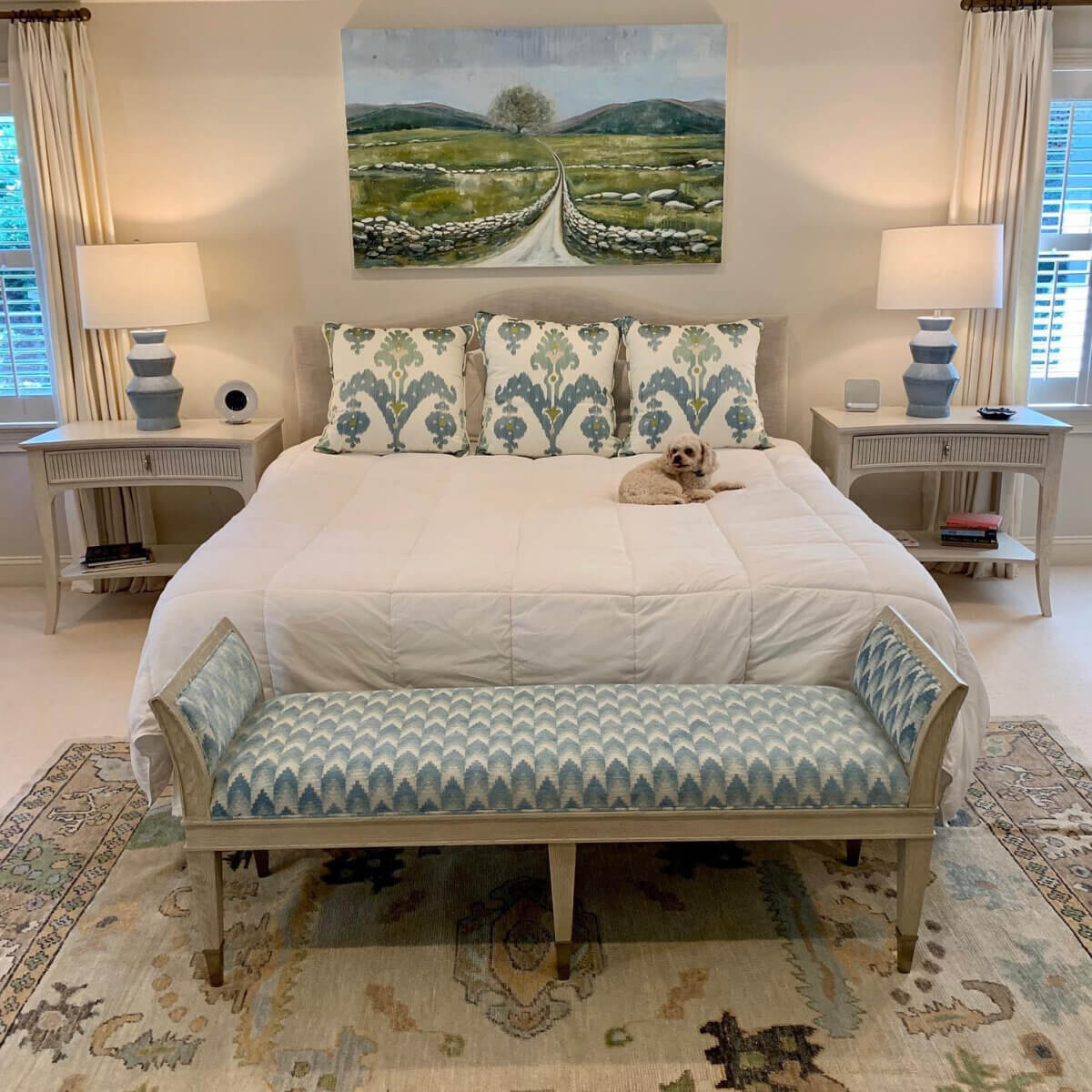 Shea Bryars on lntentional Interior Design β and Saying No to 'Amazon Living Rooms' - 5 A neatly made bed with patterned pillows, a small dog lying on top, two nightstands with lamps, a decorative bench at the foot, and a landscape painting above the bed.