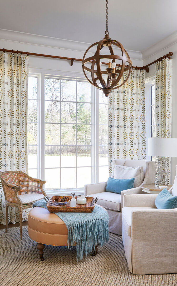 Shea Bryars on lntentional Interior Design β and Saying No to 'Amazon Living Rooms' - 3 Bright living room with large windows, patterned curtains, two beige armchairs, a cane chair, round leather ottoman, blue throw blanket, and a modern wooden chandelier.