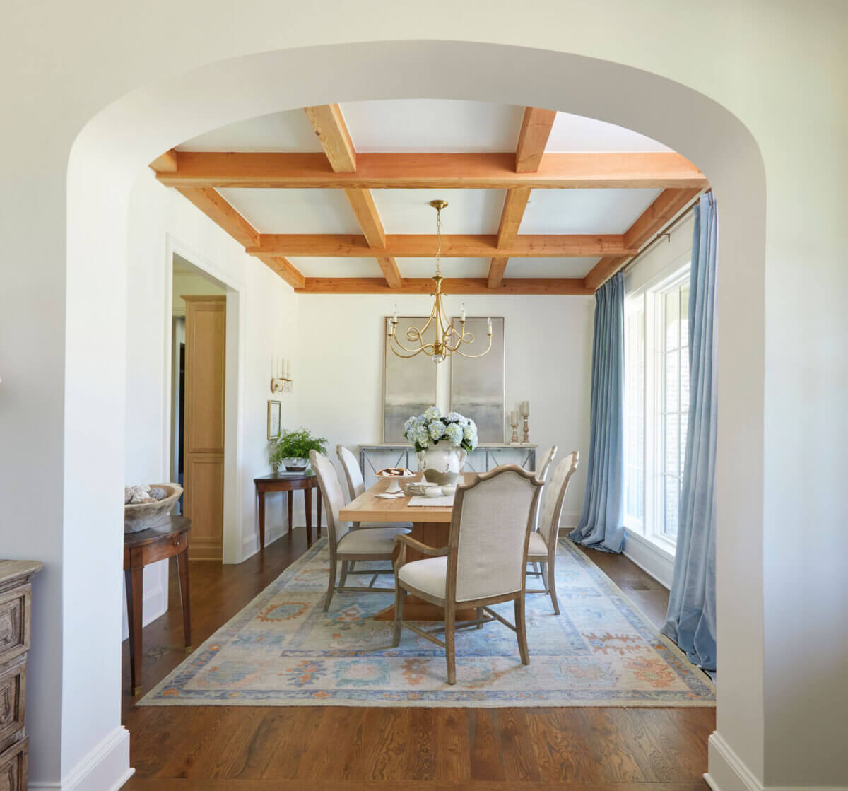 Shea Bryars on lntentional Interior Design β and Saying No to 'Amazon Living Rooms' - 12 A dining room features a wooden table with upholstered chairs, a floral centerpiece, a chandelier, large windows with blue curtains, and a beamed ceiling.