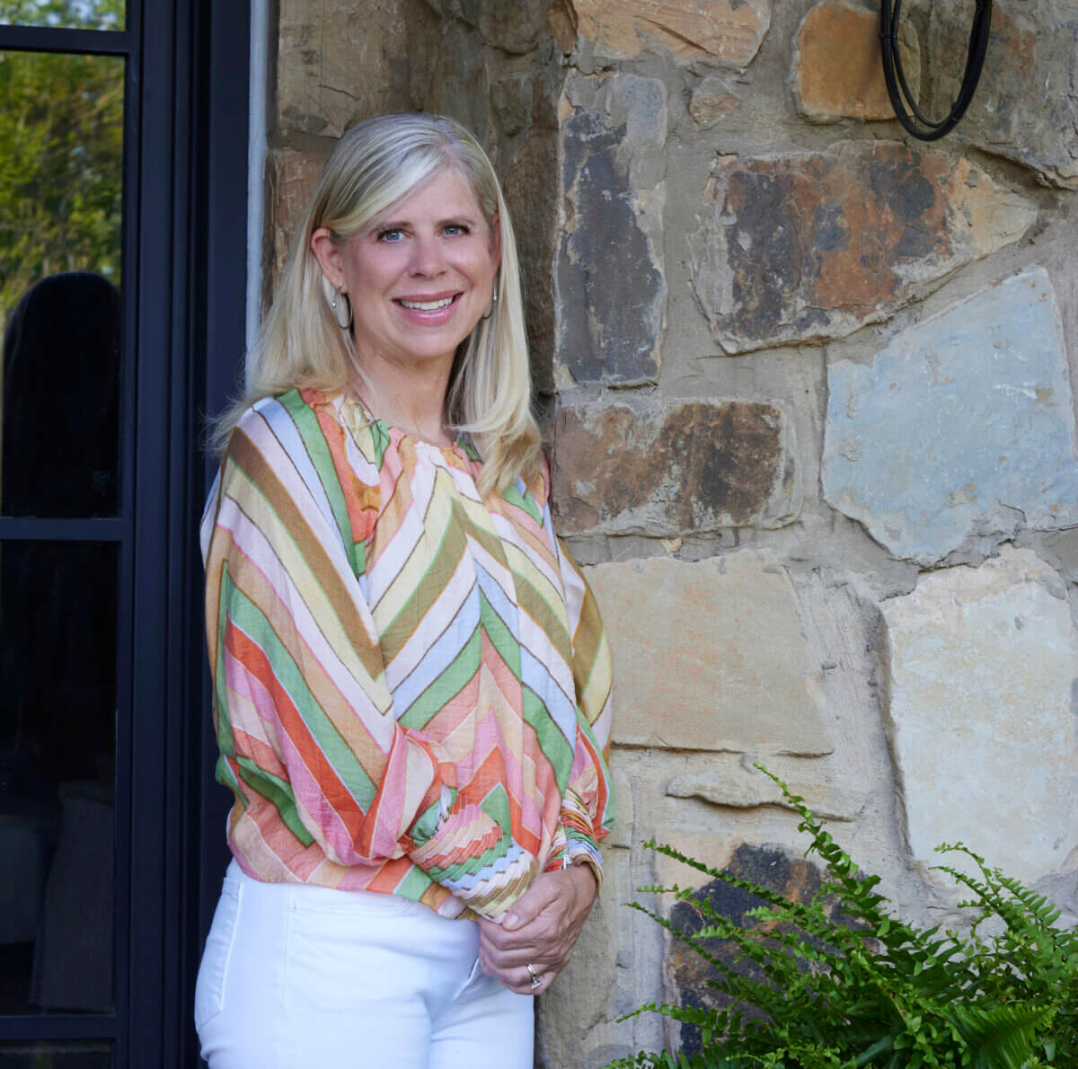 Shea Bryars on lntentional Interior Design β and Saying No to 'Amazon Living Rooms' - 2 A woman with long blond hair, wearing a colorful striped blouse and white pants, stands in front of a stone wall near a door and a fern.