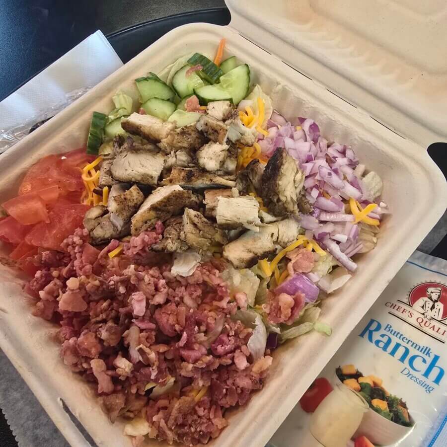 9 New Birmingham Restaurants to Try This Month - 2 A takeout salad in a compostable container with grilled chicken, chopped bacon, diced tomatoes, cucumbers, shredded cheese, and red onions, next to a packet of buttermilk ranch dressing.