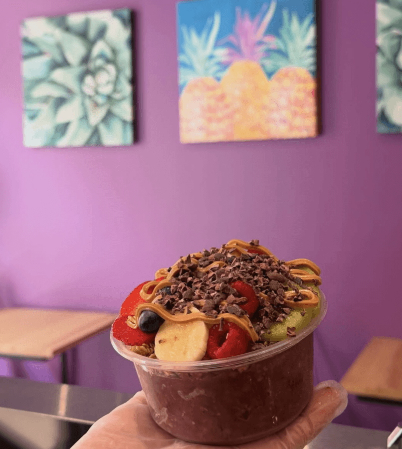 9 New Birmingham Restaurants to Try This Month - 3 A hand holds an acai bowl topped with sliced fruit and cacao nibs in front of a purple wall with tropical fruit paintings.