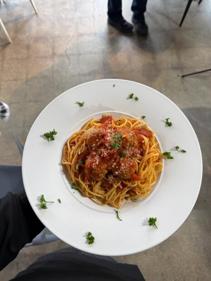 9 New Birmingham Restaurants to Try This Month - 6 A plate of spaghetti topped with tomato sauce and meatballs, garnished with grated cheese and parsley, served in one of Birmingham's new restaurants to try, held over a concrete floor with chairs and legs visible in the background.