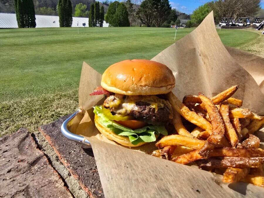 9 New Birmingham Restaurants to Try This Month - 7 A cheeseburger with lettuce, tomato, and bacon, served with fries on parchment paper, set outdoors near a golf course green—a must-try for anyone exploring top Birmingham Restaurants.