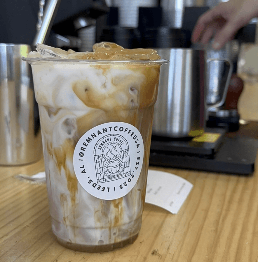 9 New Birmingham Restaurants to Try This Month - 10 A plastic cup of iced coffee with milk swirls sits on a wooden counter. A metal straw and a Remnant Coffee USA label are visible, reflecting the creativity seen in new restaurant concepts at top Birmingham restaurants. Barista equipment is in the background.