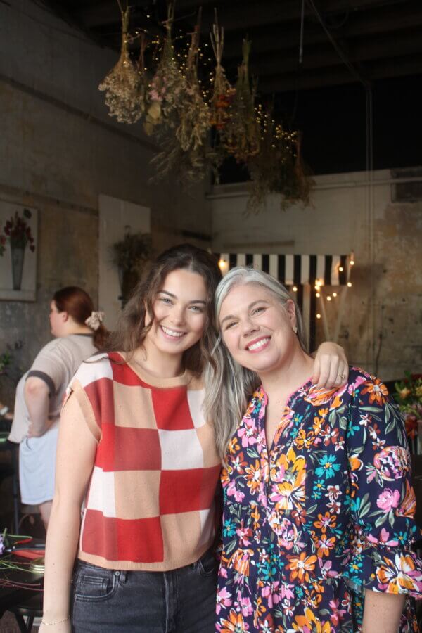 Meet the Mother-Daughter Duo Behind Wild Honey Flower Truck - 6 Two women, a mother-daughter duo, smile and pose together indoors with dried flowers and string lights hanging above. Other people are visible working in the room, capturing the warm spirit of Wild Honey Flower Truck.
