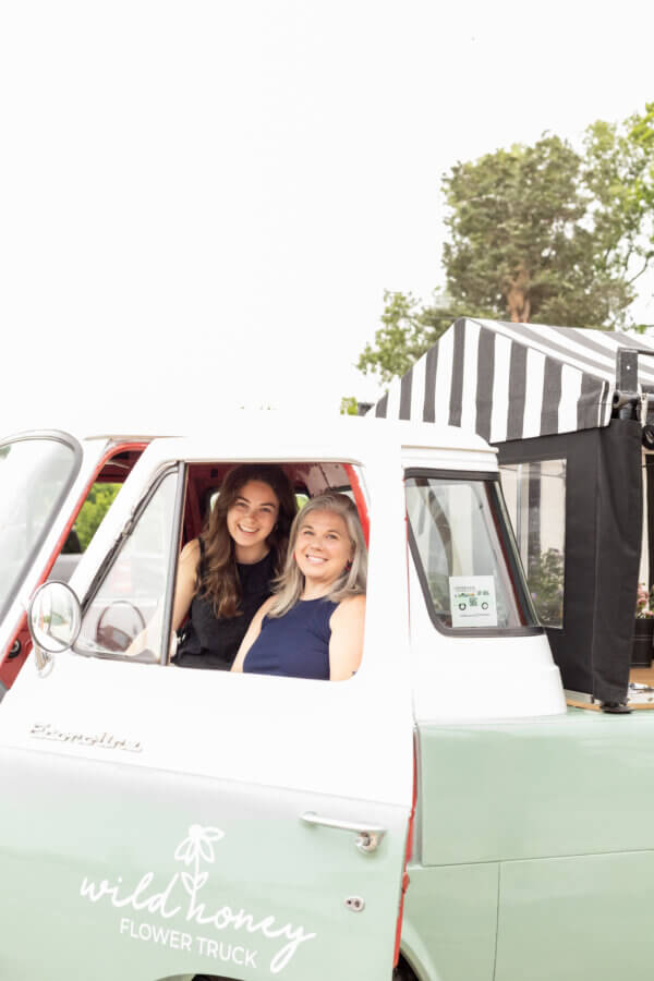 Meet the Mother-Daughter Duo Behind Wild Honey Flower Truck - 2 A smiling mother-daughter duo sits in the front seat of a vintage Wild Honey Flower Truck, with an outdoor market scene unfolding in the background.