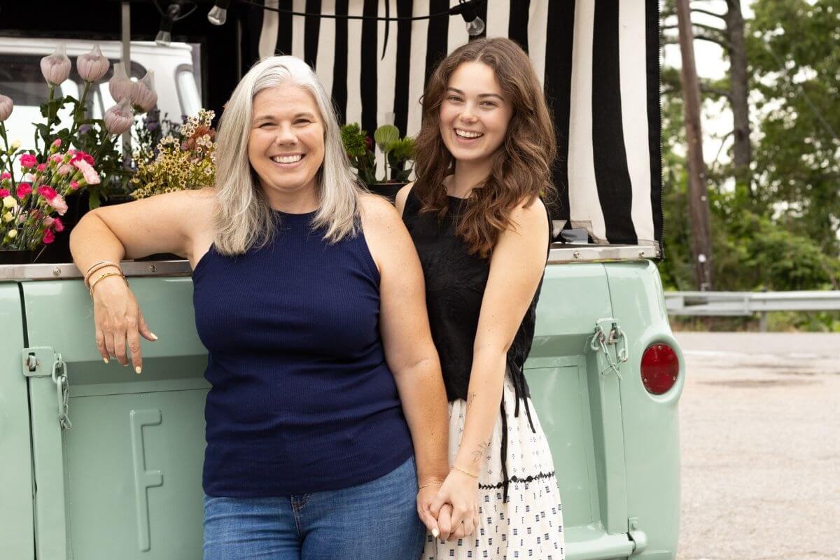 Two women stand and smile in front of a vintage green flower truck with flowers displayed in the back. One woman is older with gray hair, while the other is younger with brown hair.