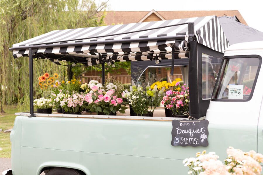 Meet the Mother-Daughter Duo Behind Wild Honey Flower Truck - 3 A mint green flower truck, Wild Honey Flower Truck, with a black-and-white striped awning displays fresh bouquets. A chalkboard sign reads "Build a Bouquet: 5 Stems." This charming setup is run by a creative mother-daughter duo.