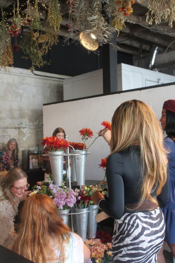 Meet the Mother-Daughter Duo Behind Wild Honey Flower Truck - 4 People gather around flower arrangements on a table in a rustic indoor setting with hanging dried plants overhead, curated by the mother-daughter duo behind Wild Honey Flower Truck.