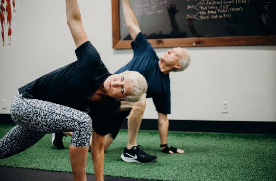 Two older adults wearing exercise clothes perform a stretching exercise on a green mat in a gym, guided by EIM Trainer Cindy Pounds, with a chalkboard and anatomical chart in the background.