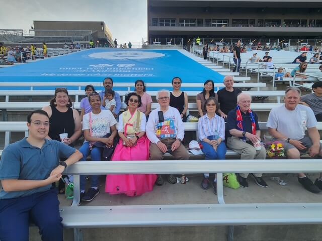 From War Refugee to Biochemist to ... Tap Dancer! Meet Helen Kim - 3 A group of people sit on bleachers at an outdoor stadium, some in casual clothes and others in traditional attire, reflecting Unity in Diversity as seen in Asian cultures. The stadium appears partially filled.