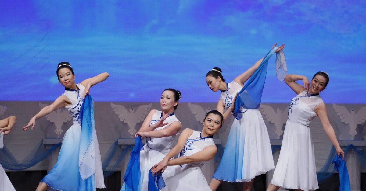 From War Refugee to Biochemist to ... Tap Dancer! Meet Helen Kim - 4 Five women in white and blue dresses perform a synchronized dance on stage against a blue backdrop, celebrating Asian cultures and showcasing Unity in Diversity.