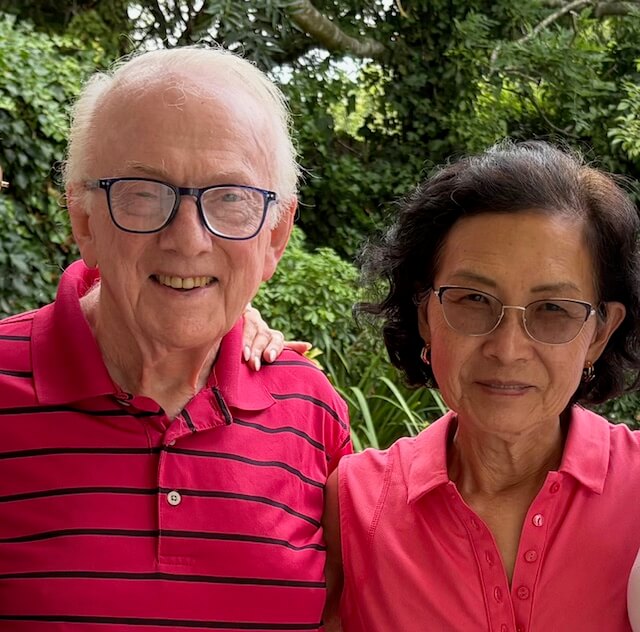 From War Refugee to Biochemist to ... Tap Dancer! Meet Helen Kim - 2 Helen Kim stands side by side by her husband, Stephen Barnes, smiling softly with greenery in the background.