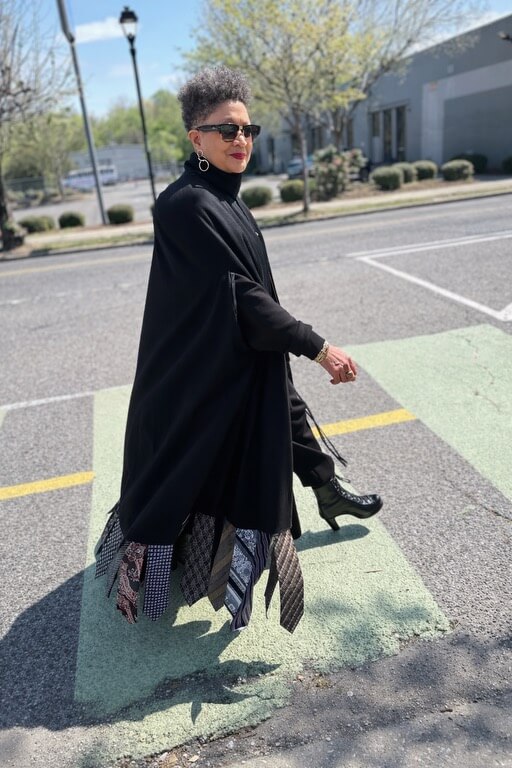 She Curates One of Birmingham’s Top Vintage & Consignment Boutiques - 3 A woman wearing sunglasses, a black coat, and a skirt with patterned ties—perhaps curated from Birmingham vintage boutiques—walks across a crosswalk on a sunny day.